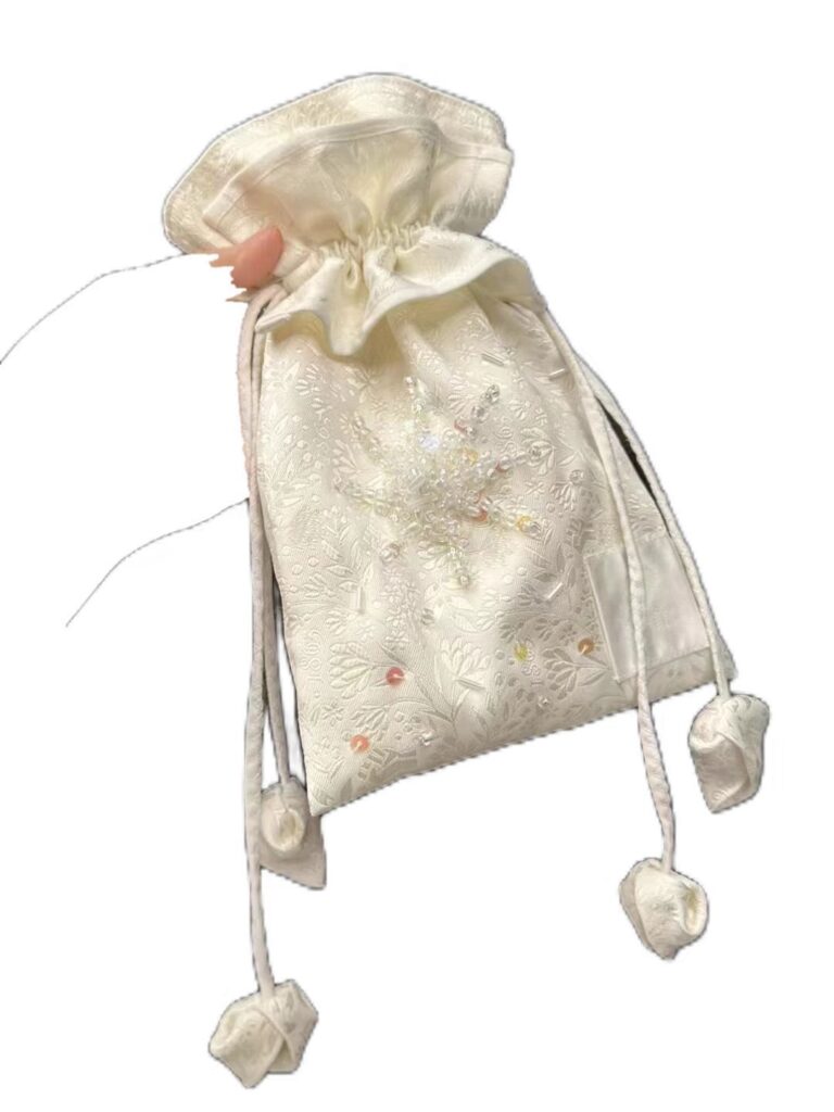 Bolso de tela bordada en color marfil, decorado con motivos florales y detalles brillantes, con cordón ajustable.